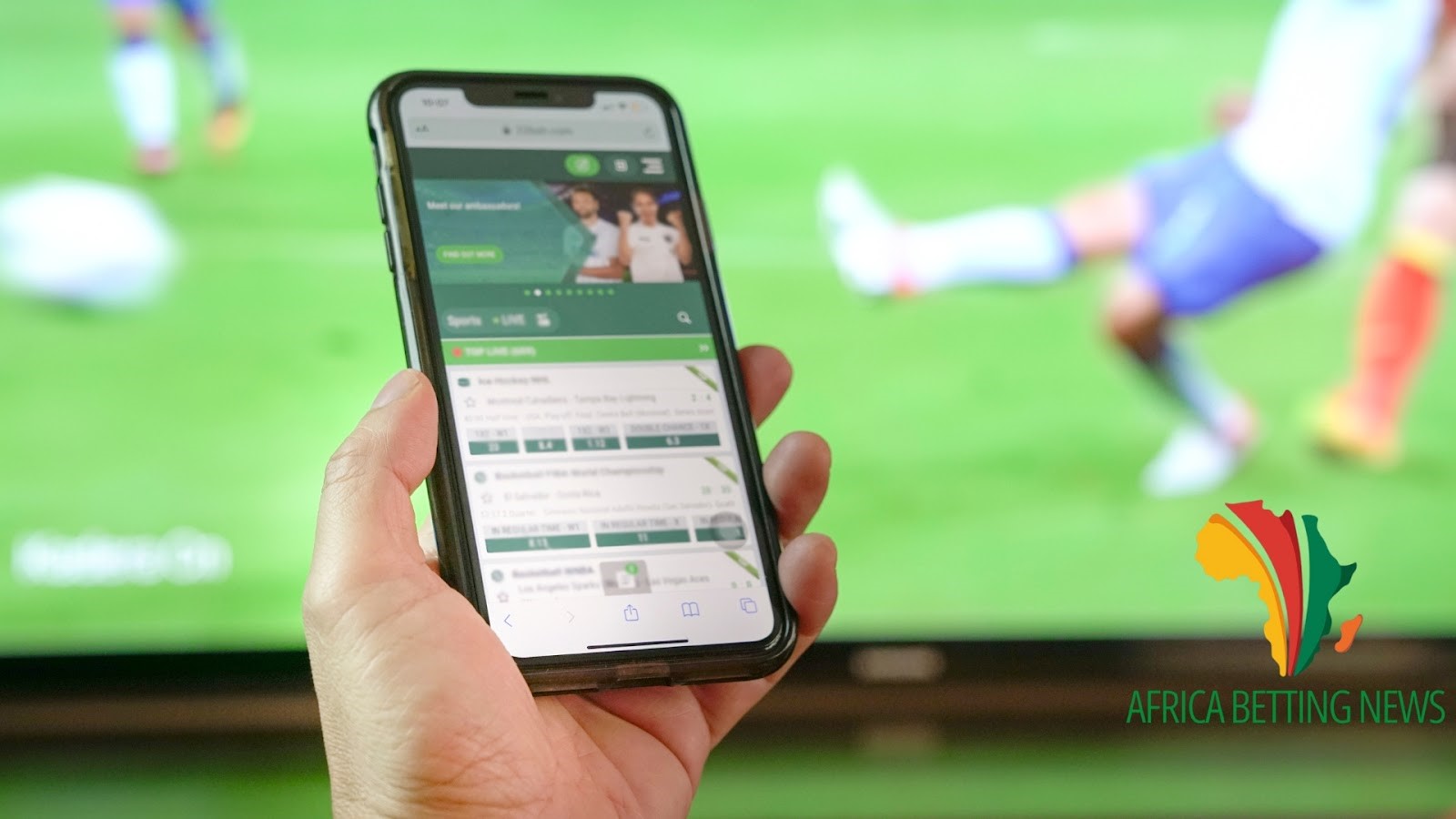 A person holding a smartphone with a betting app open on the screen. In the background, a blurred image of a soccer match is visible on a TV screen. The Africa Betting News logo is displayed in the lower right corner of the image.
